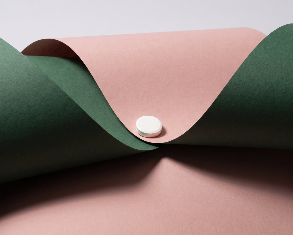 A single white dissolving tablet resting in a fold of forest-green and blush paper